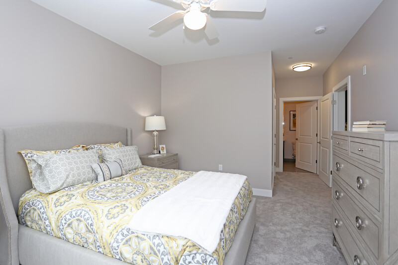 Staged primary bedroom with upholstered queen bed, patterned bedding, dresser, and ceiling fan, with view into ensuite bathroom