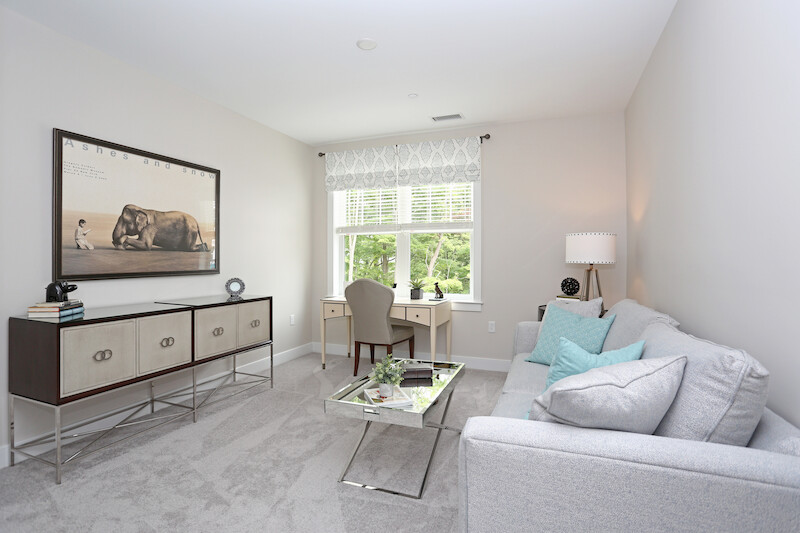 Versatile den styled as a home office with a writing desk, accent sofa, custom cabinetry, and natural light from a tree-line view