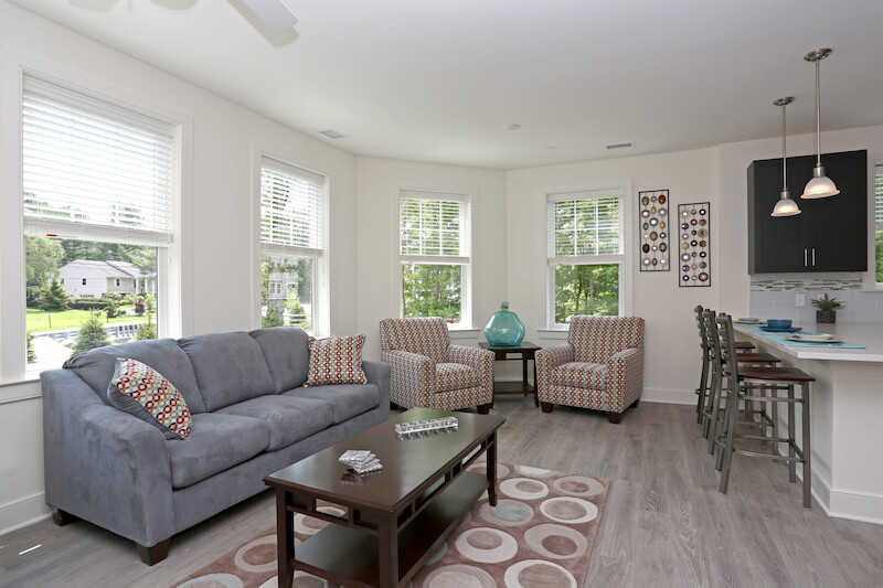 Bright living room with bay windows, gray sofa, accent chairs, wide-plank flooring, and open sightline to the kitchen breakfast bar